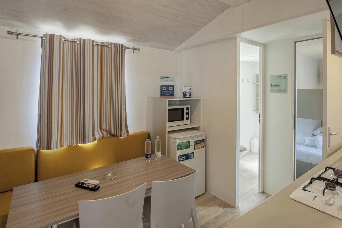 A compact kitchen with a microwave, a small refrigerator, and a gas stove. There is a dining table with chairs and a yellow sofa.