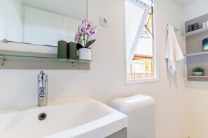 Een badkamer met een witte wastafel, een spiegel, een raam met uitzicht en een plank met toiletartikelen.