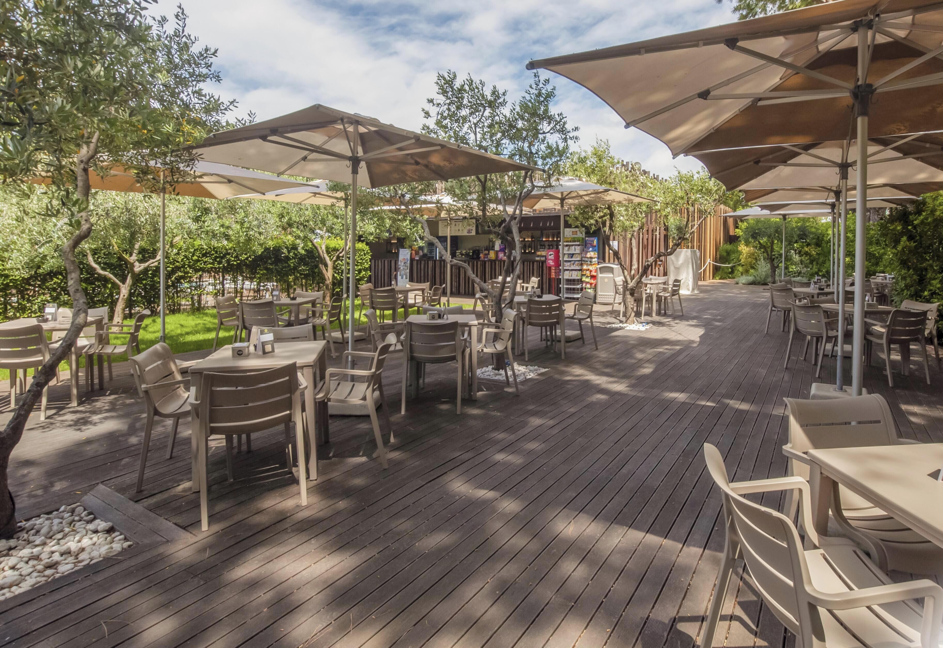 Un espace extérieur aménagé en terrasse, avec des tables et des chaises placées sous de grands parasols, le tout entouré de verdure et d’arbres.