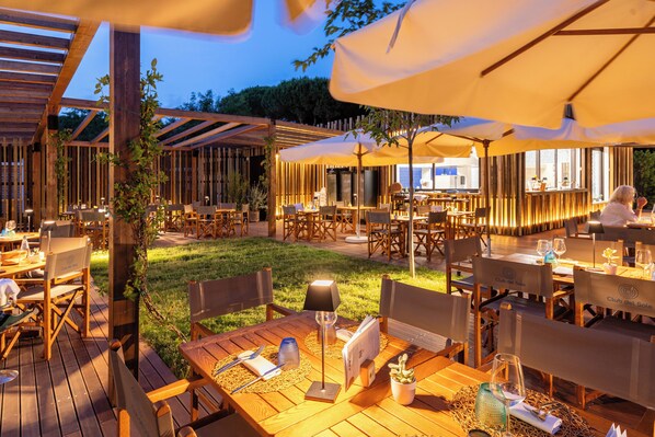Een buiten eethoek met houten tafels en stoelen, parasols en een sfeervolle verlichting.