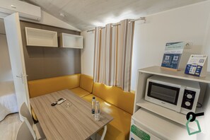 Una habitación compacta con una mesa de madera, un banco acolchado amarillo, un microondas y un refrigerador pequeño.