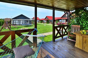 A wooden deck with a hammock, a wooden table with fruit, and a view of a green lawn and buildings in the background. - Single-story Holiday Homes With Terraces (Darlowo)