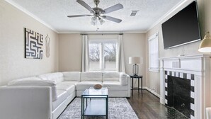 A living room with a white sofa, a glass coffee table, a fireplace, and a ceiling fan. - Stylish & Modern Private Getaway in Richardson! (Richardson)