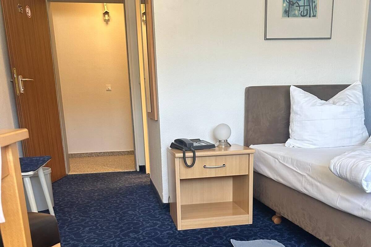 A hotel room with a bed, a desk with a telephone, a small nightstand, a chair, and a carpeted floor.