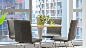 Ein modernes Interieur mit einem runden Tisch und vier grauen Stühlen, einer Topfpflanze auf dem Tisch sowie einem Blick auf eine Stadtlandschaft durch große Fenster.