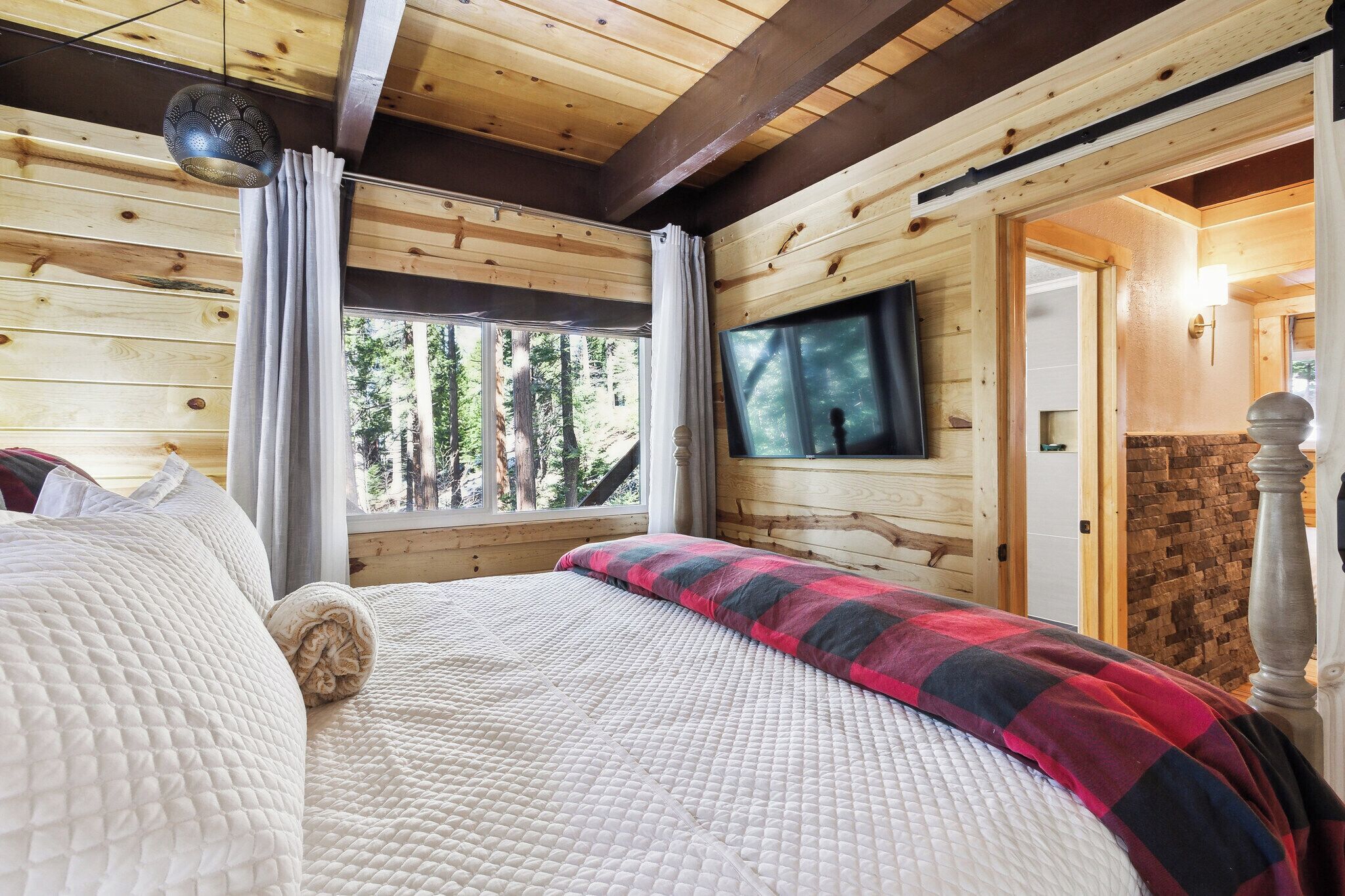 A wooden cabin bedroom with a bed, curtains, a TV, and a stone wall.