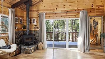 A wooden cabin interior with a stone fireplace, a hanging chair, and a balcony with a view of trees.