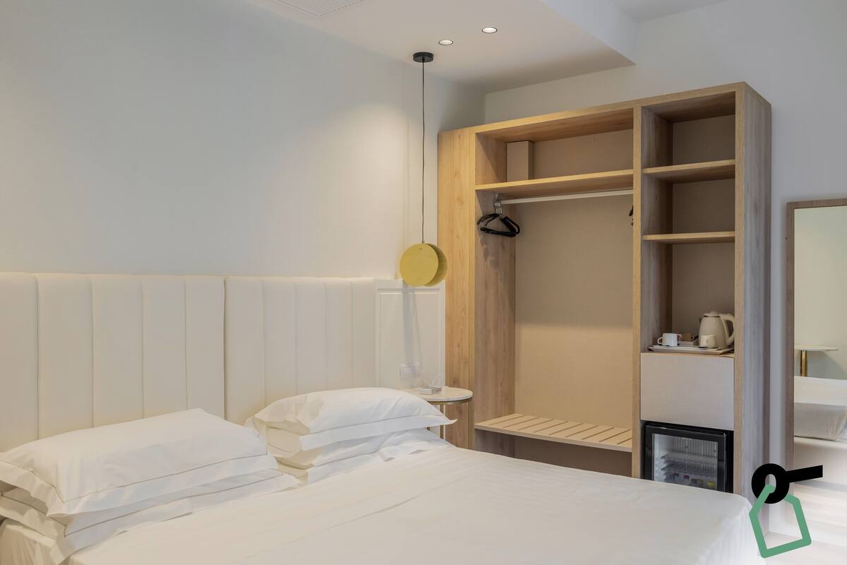 Une chambre d’hôtel avec un lit, une armoire en bois, une petite table avec une bouilloire et un luminaire suspendu.