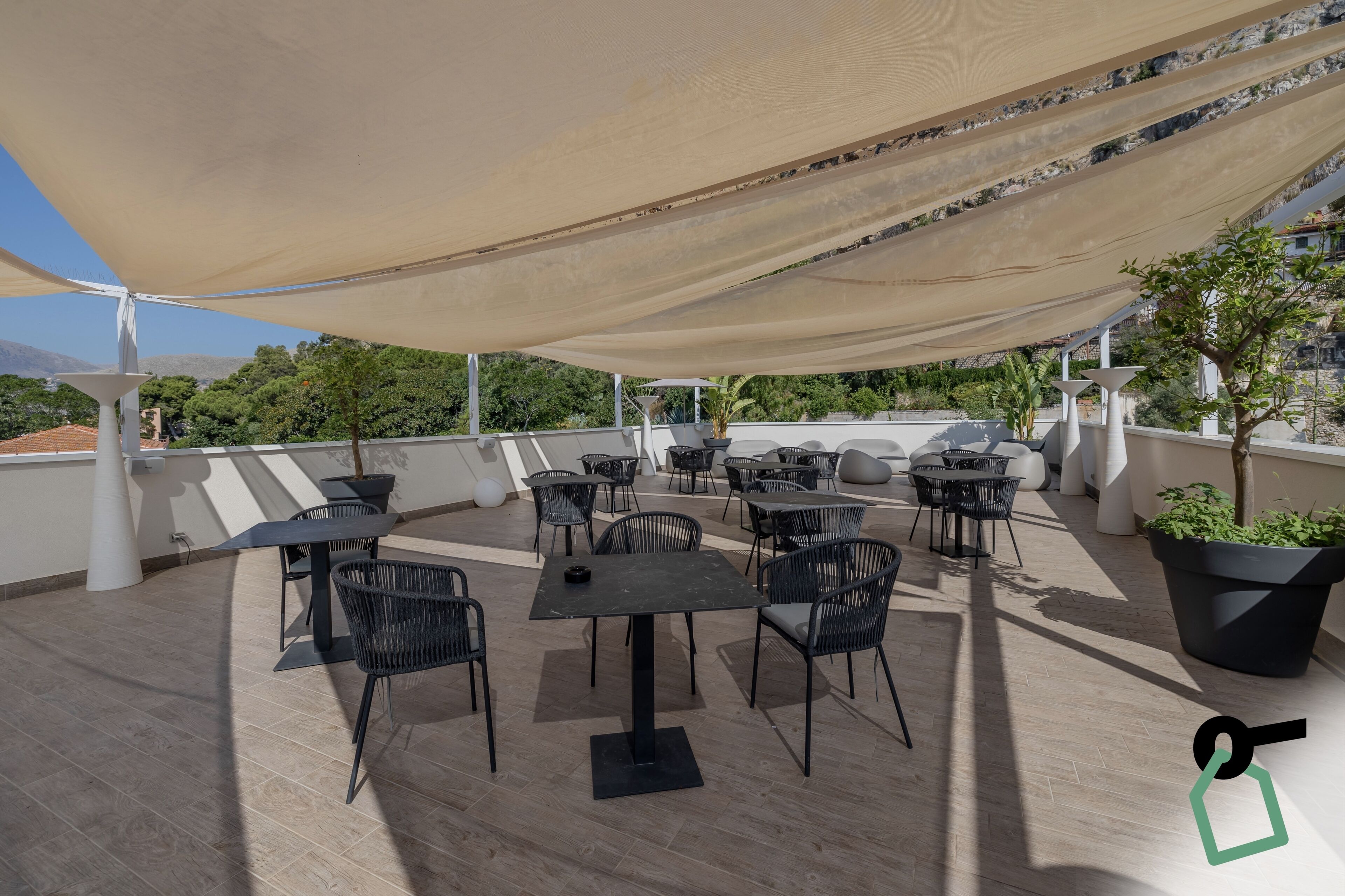 An outdoor terrace with multiple tables and chairs, a large canopy providing shade, and potted plants.