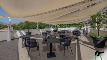 An outdoor terrace with multiple tables and chairs, a large canopy providing shade, and potted plants.