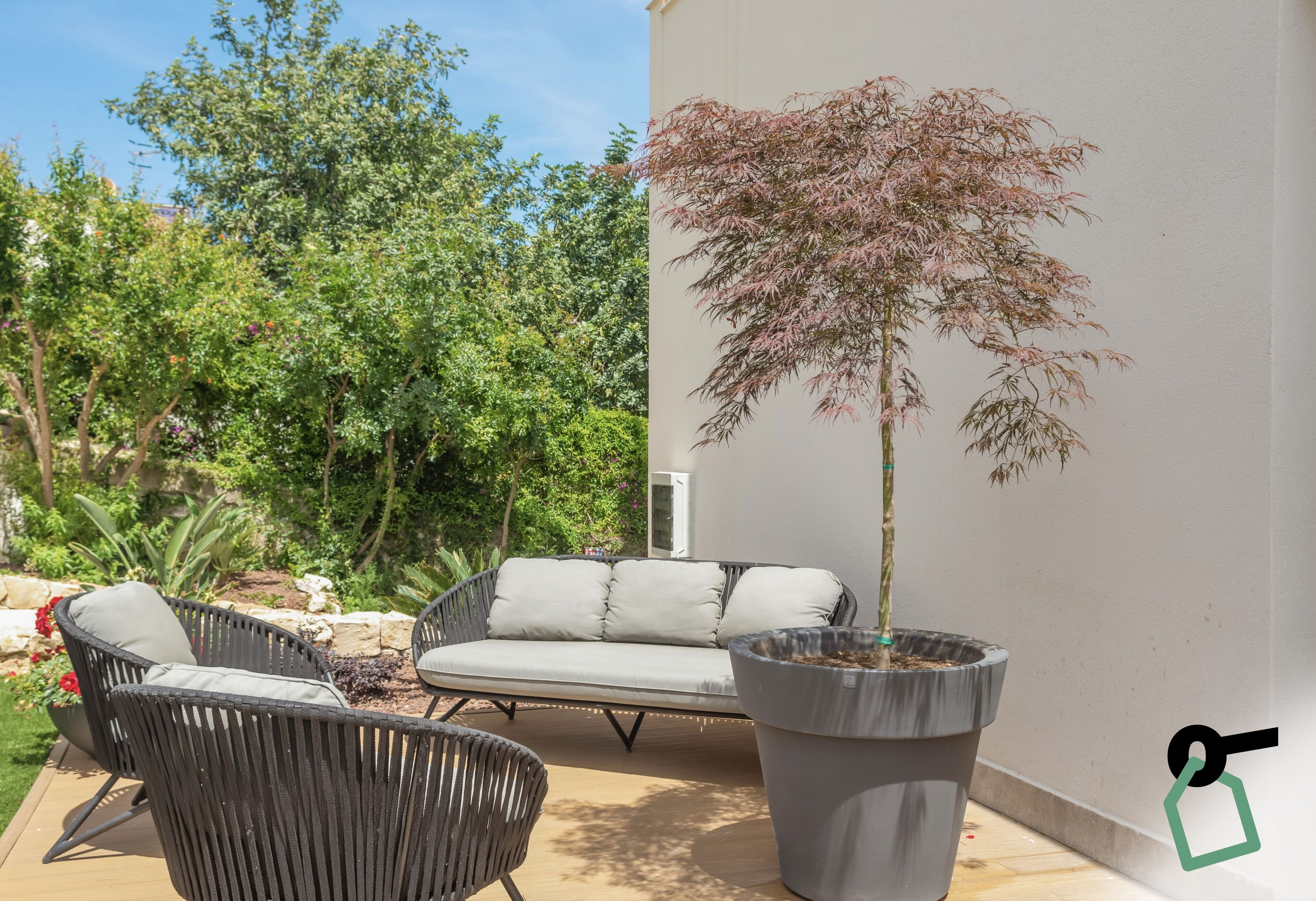 Un área de estar al aire libre con sillas de mimbre y un sofá blanco, un árbol grande en maceta y una pared blanca.