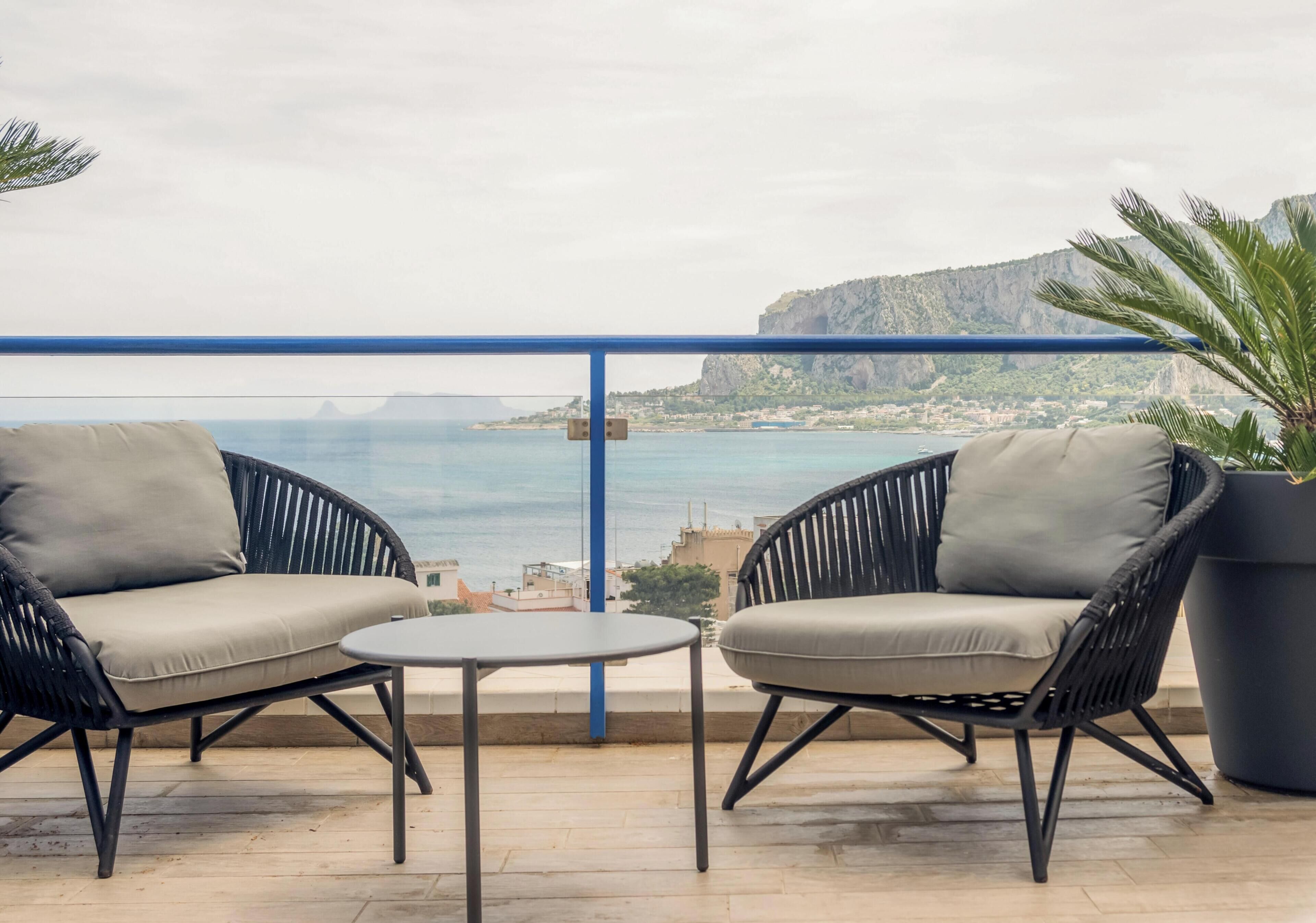 Un balcón con dos sillones de mimbre negro y una mesita, con vistas a la costa y una montaña al fondo.
