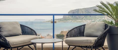 A balcony with two chairs and a small table, overlooking a coastal view with buildings and cliffs.