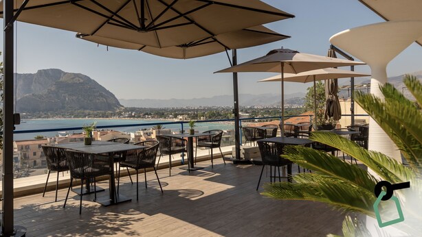 An outdoor terrace with tables and chairs, umbrellas, and a view of the sea and mountains. - Hotiday Mondello Beach (Palermo)