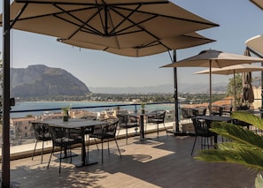 An outdoor terrace with tables and chairs, overlooking a coastal view with mountains in the distance. - Hotiday Mondello Beach (Palermo)