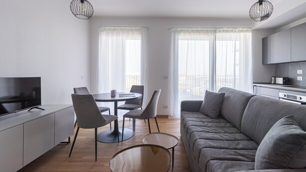 A modern living room with a grey sofa, a dining table with chairs, a TV, and a kitchen area with a microwave. - Italianway Comfort - Pasolini 70 B - Feel Uptown (Milano)