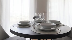 A round dining table set with white plates, bowls, and clear glasses, positioned near a window with sheer curtains. - Italianway Comfort - Pasolini 70 B - Feel Uptown (Milano)