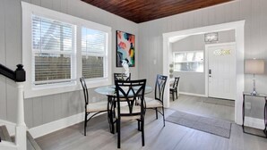 A dining area with a wooden ceiling, white walls, and a large window with blinds. There is a table with chairs and a painting on the wall.