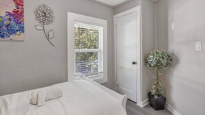 A bedroom with a bed, a window with blinds, a door, a wall-mounted artwork, and a potted plant.