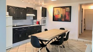A compact kitchen with dark cabinets, a white refrigerator, a microwave, and a coffee maker. There is a dining table with four chairs and a large abstract painting on the wall. - Gîte des Orgues d'Ille Eucalyptus (Ille-sur-Têt)