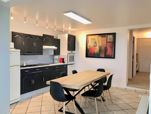 Una cocina compacta con gabinetes oscuros, un refrigerador blanco, un microondas y una cafetera. Hay una mesa de comedor con cuatro sillas y un cuadro abstracto grande en la pared.