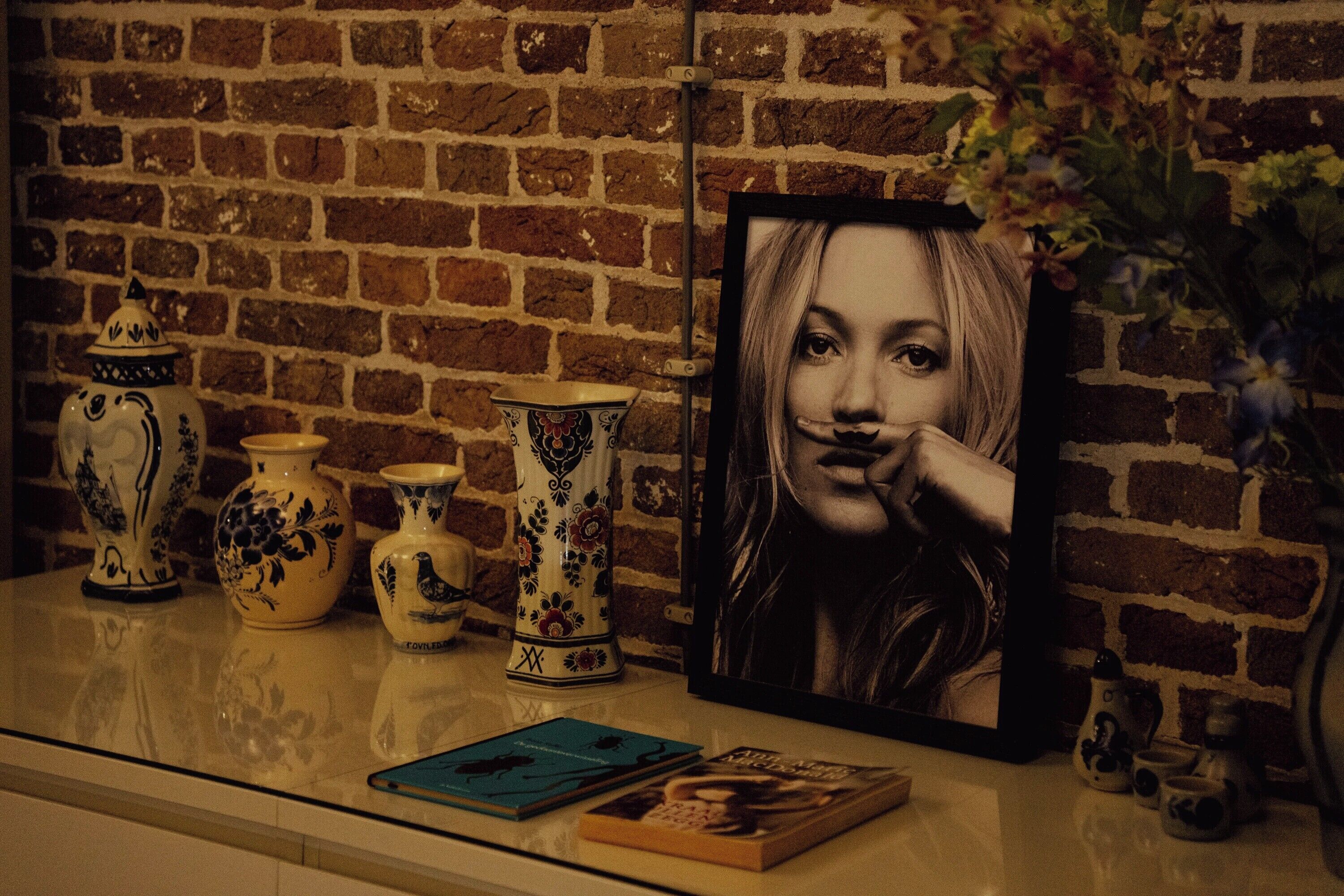 A brick wall, a framed portrait, and decorative vases on a shelf.