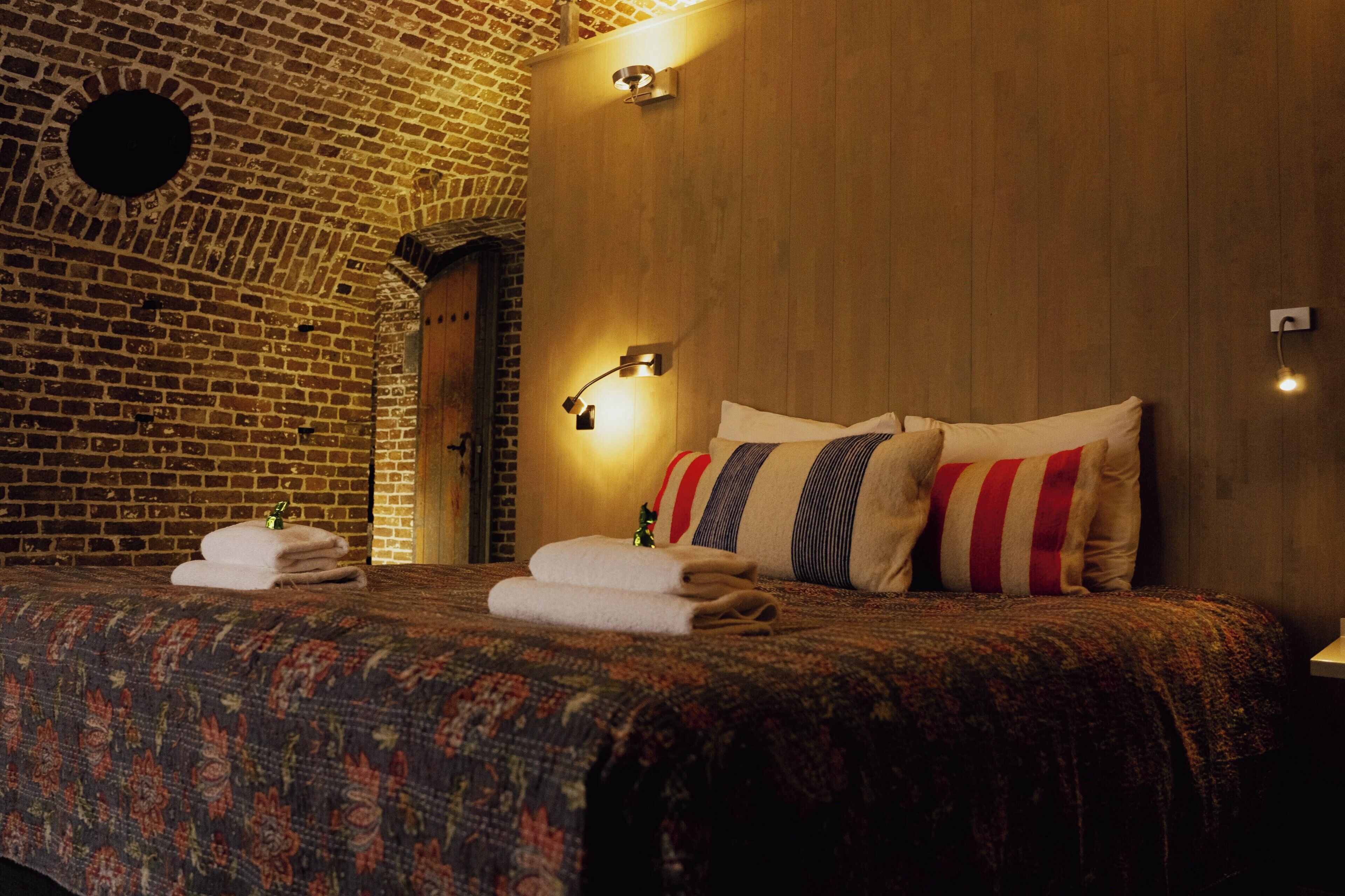 Une chambre d’hôtel avec un mur en briques, un lambris en bois, un lit recouvert d’une courtepointe à motifs, deux oreillers et une table de chevet avec une lampe.