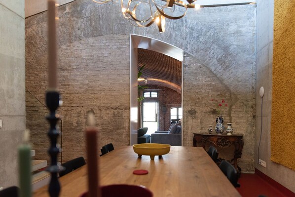 A dining area with a wooden table, chairs, and a chandelier. There is a brick wall and a view of another room with a sofa and a lamp.