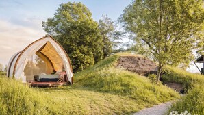 A tented accommodation with a bed, a small table, and a chair, surrounded by grassy terrain and trees. - Fort Vuren (Vuren)