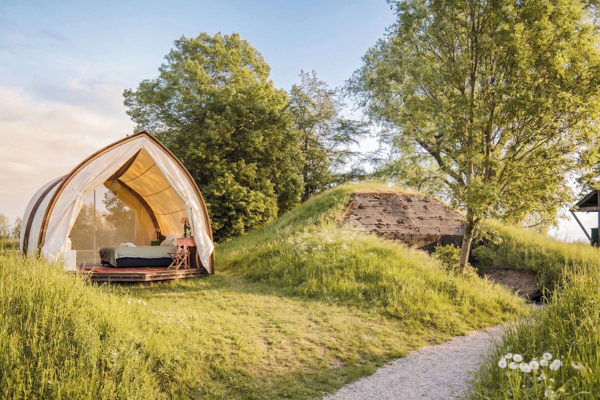A tented accommodation with a bed, a small table, and a chair, surrounded by grassy terrain and trees.