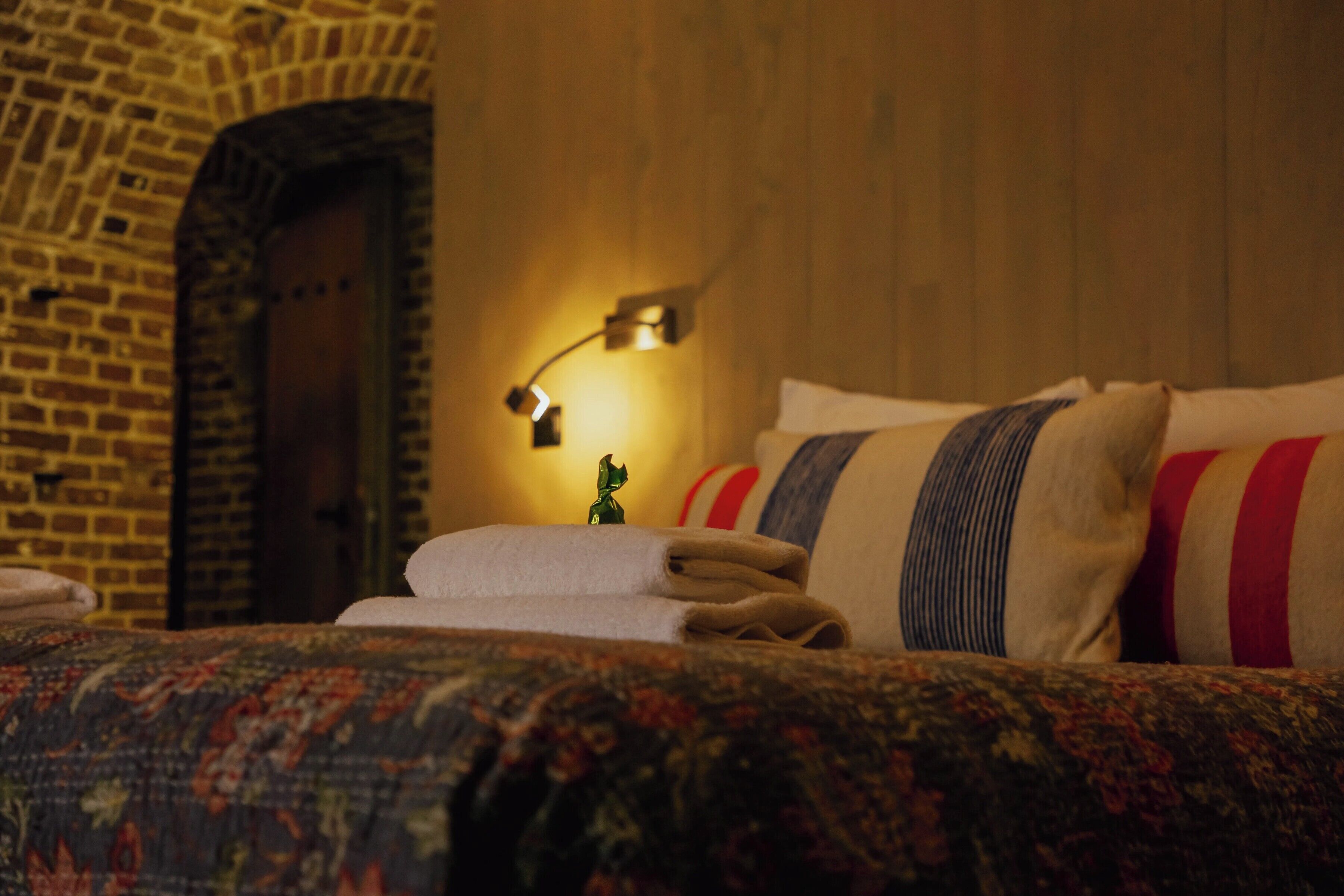 A hotel room with a brick wall, a wooden door, a bed with a patterned bedspread, and a nightstand with a lamp.
