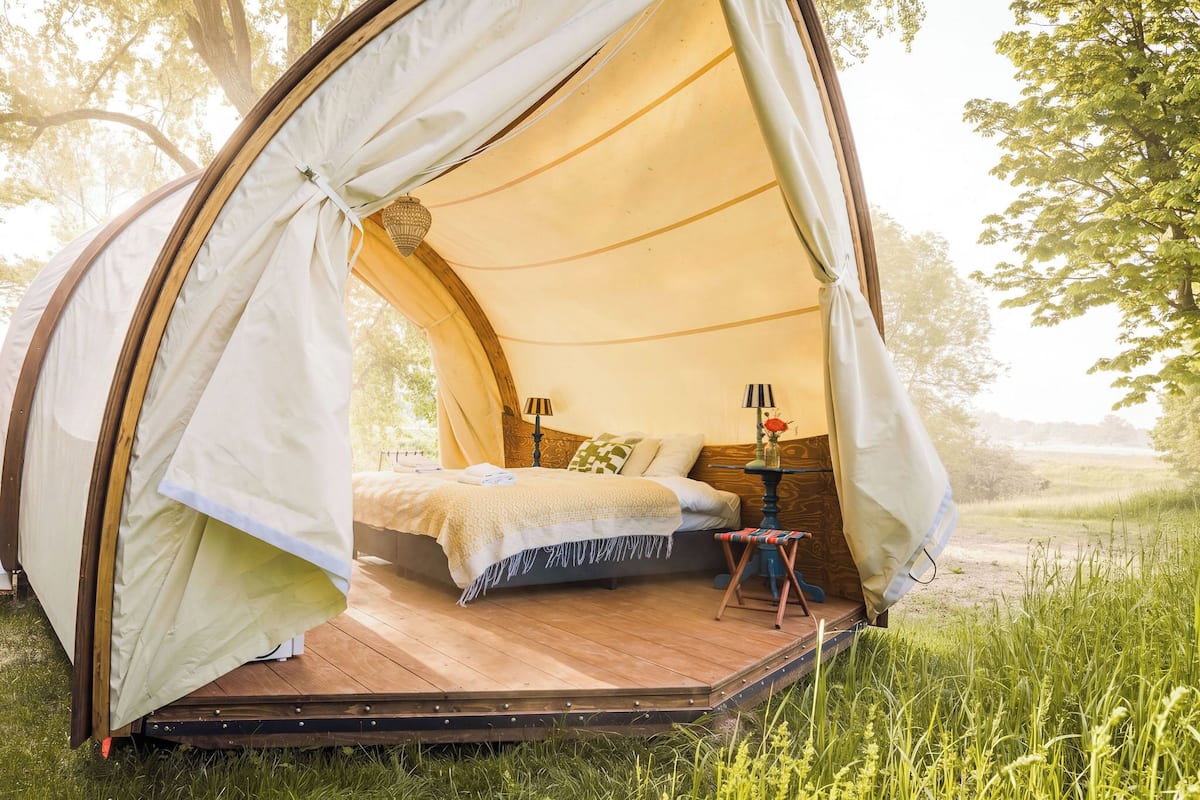 A cozy tent with a wooden platform, a bed with pillows, a small table with a lamp, and a vase with flowers.