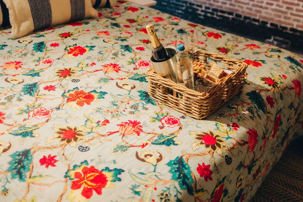 A bed with a floral bedspread, a wicker basket with bottles, and a brick wall with a small window.