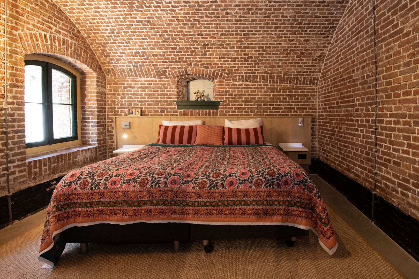 Una habitación con pared de ladrillo, cama con colcha estampada y ventana con vistas.