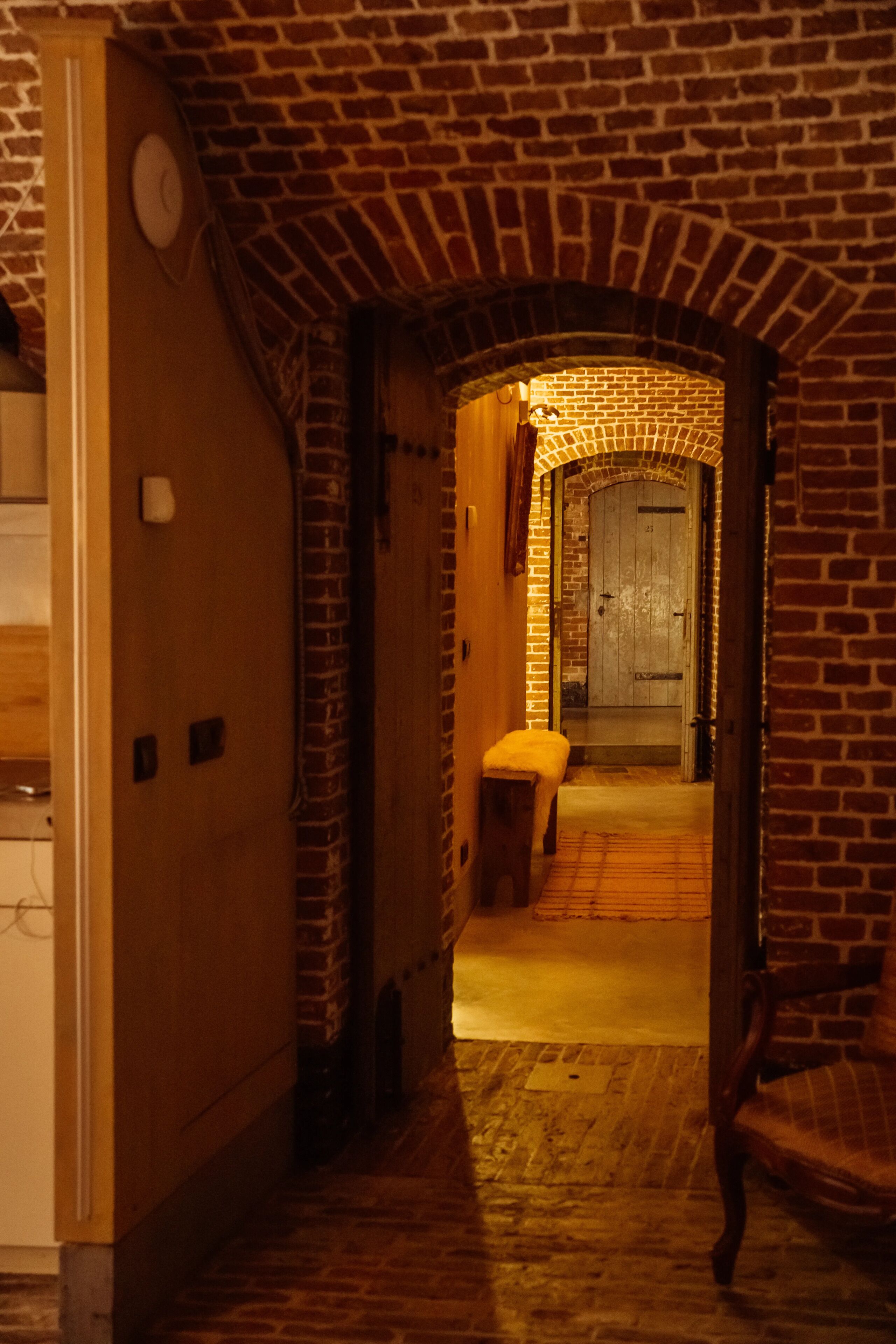 A narrow hallway with a brick archway leading to a door. A bench is placed against the wall, and a chair is visible to the right.
