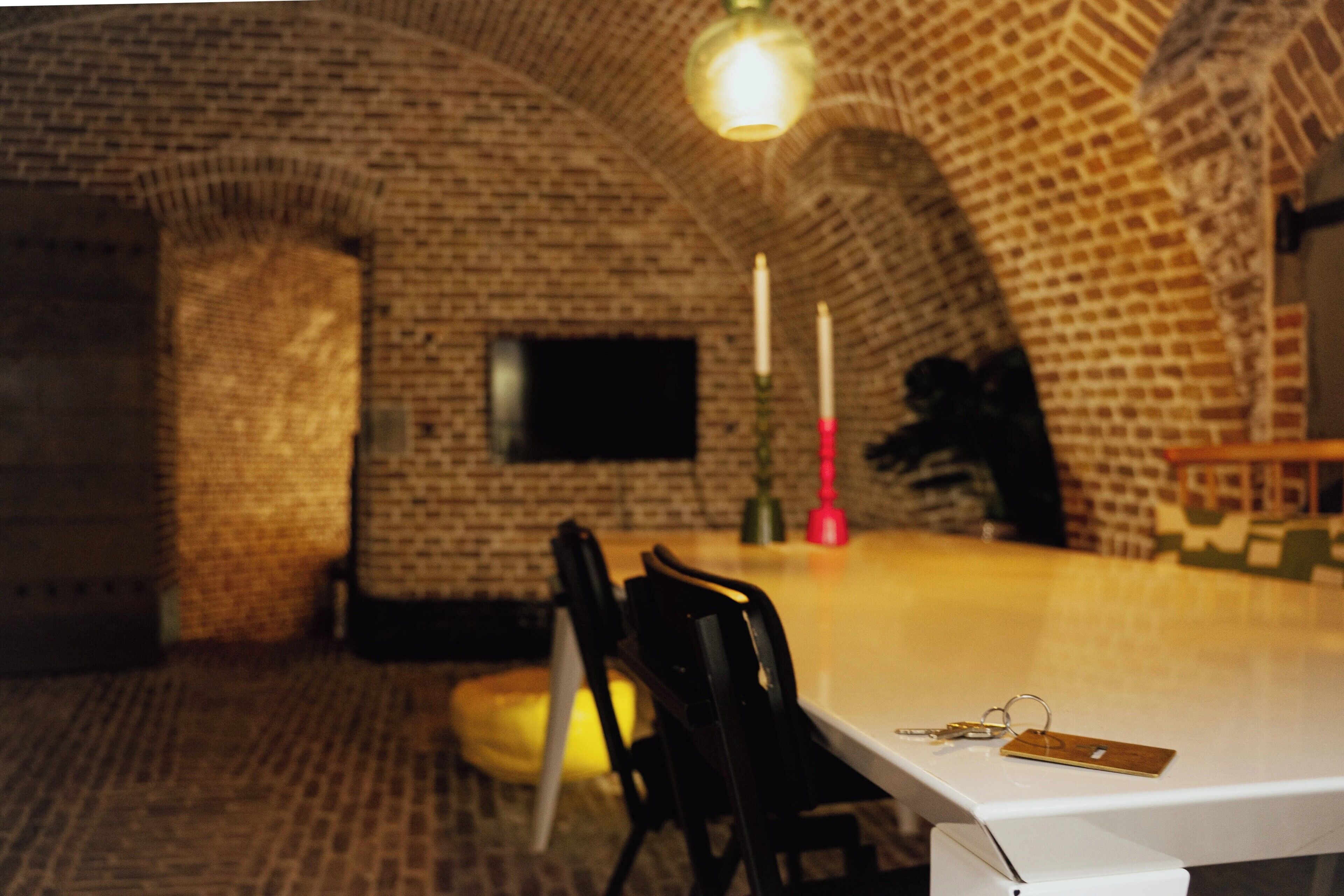 Una habitación con pared de ladrillo, una mesa blanca, sillas negras y un televisor montado en la pared.