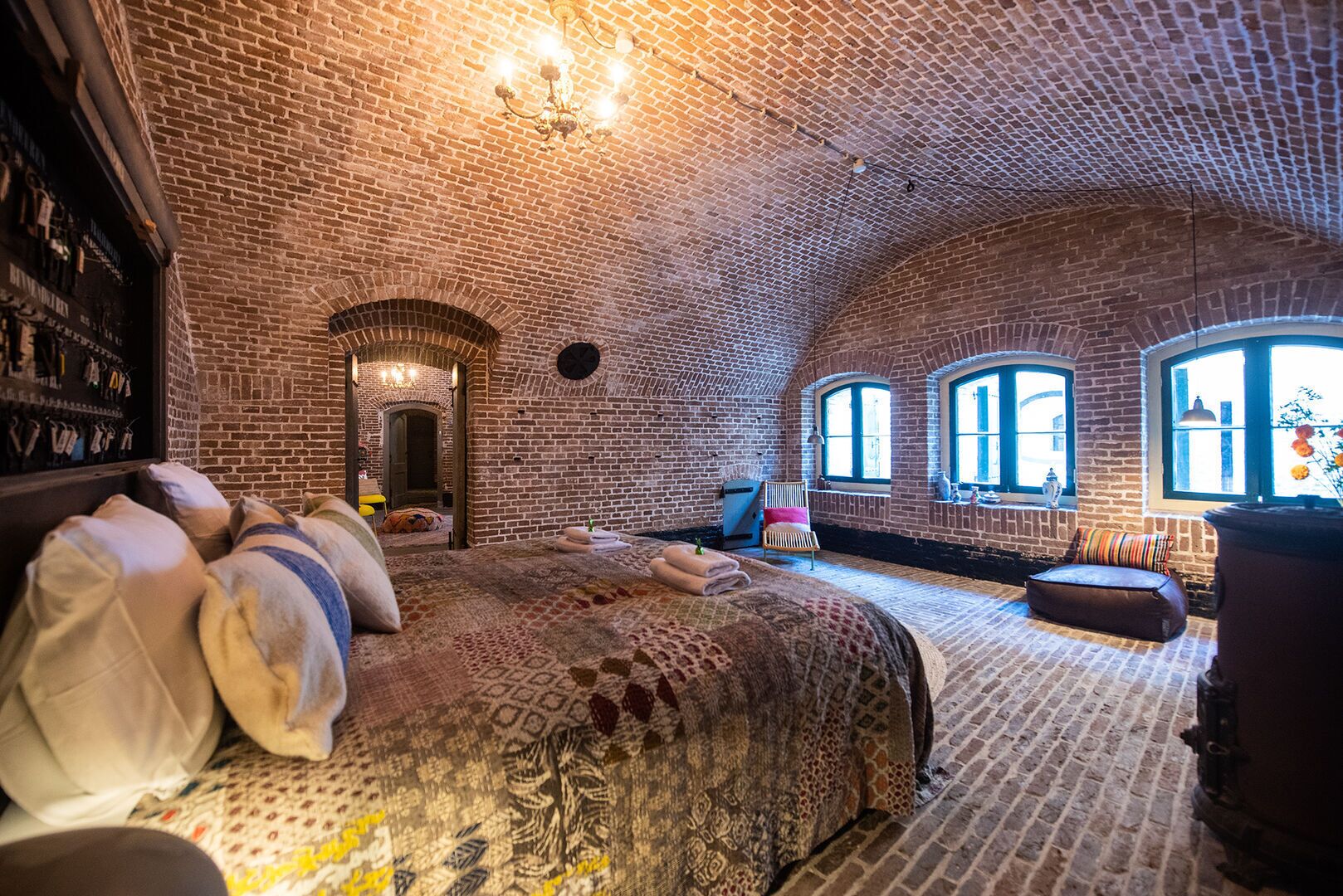 Een kamer met met bakstenen muren, een groot bed met beddengoed met een patroon, een houten stoel en een kleine tafel. Drie ramen zorgen voor natuurlijk licht.