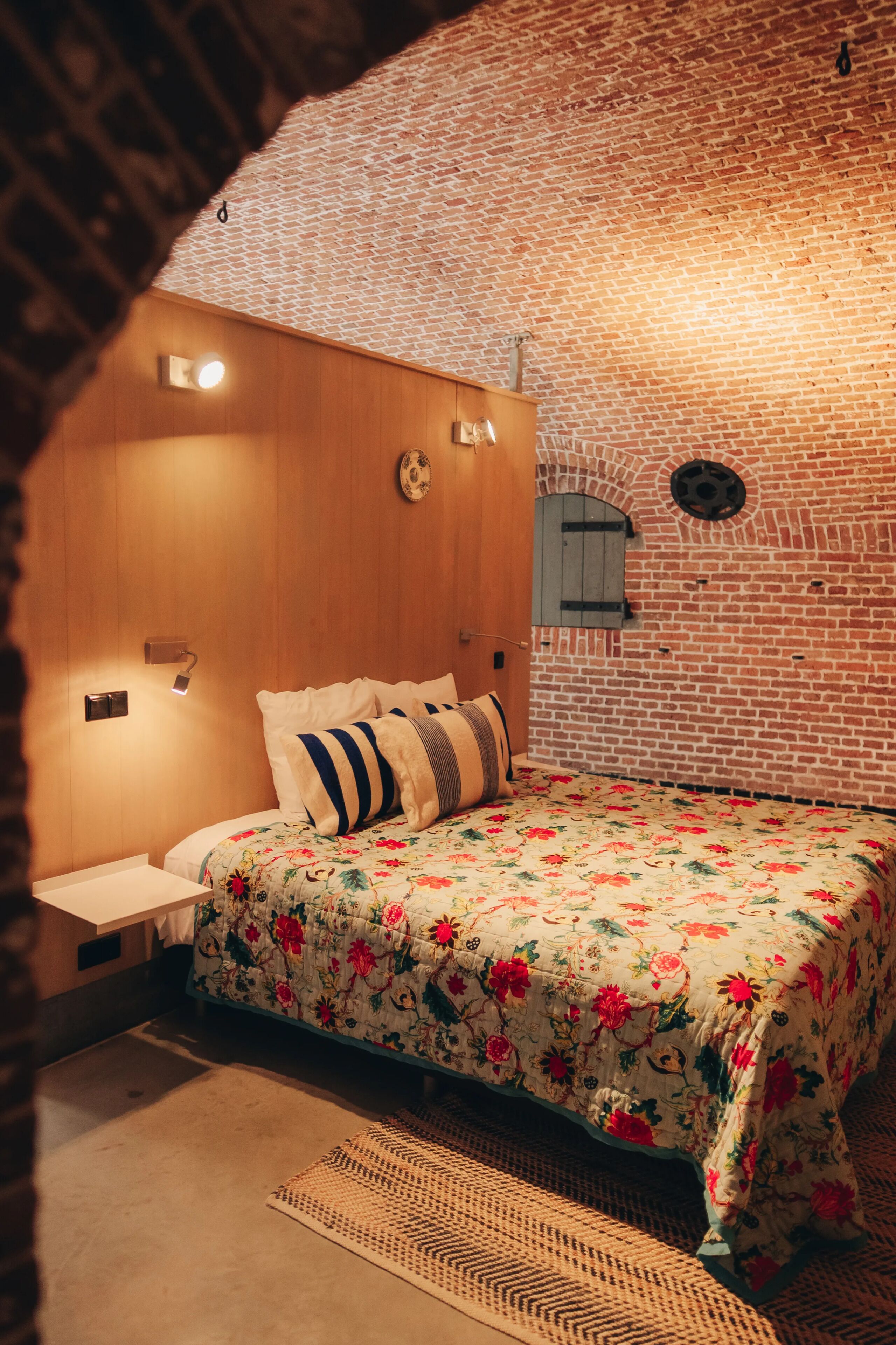 Una habitación con pared de ladrillo, cabecero de madera, colcha estampada y alfombra tejida.