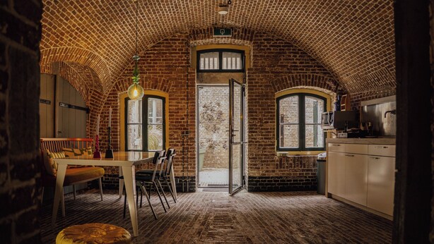 A rustic interior with a brick ceiling, a dining area with a table and chairs, a kitchen with cabinets and a stove, and a large arched doorway. - Fort Vuren (Vuren)