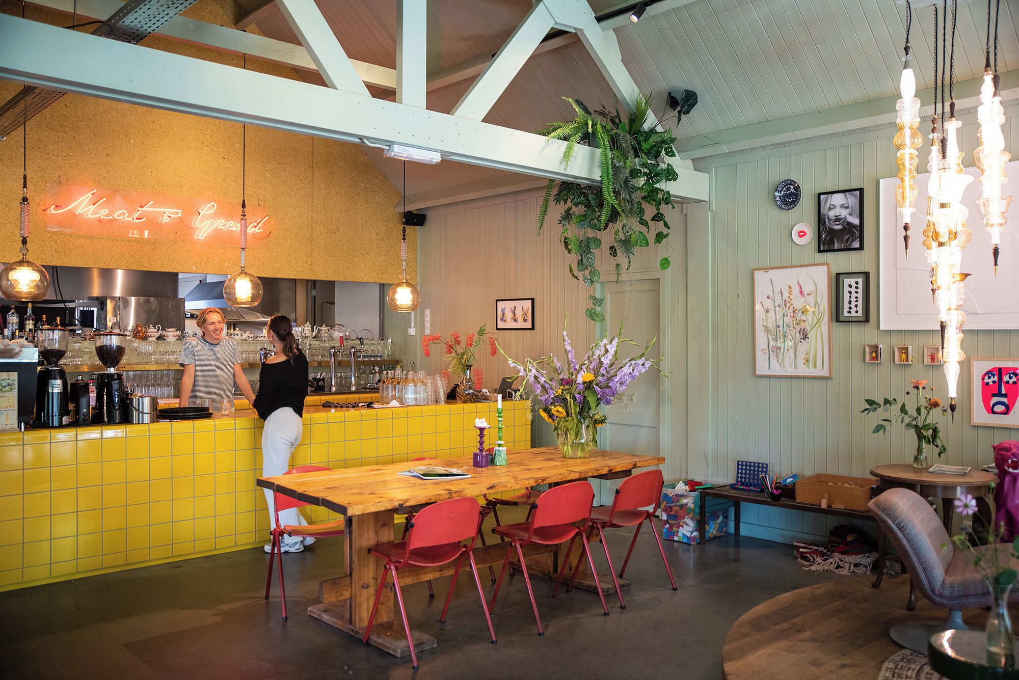 Un ristorante con zona bar, tavoli in legno, sedie rosse, piante pensili e diverse opere d'arte incorniciate alle pareti.