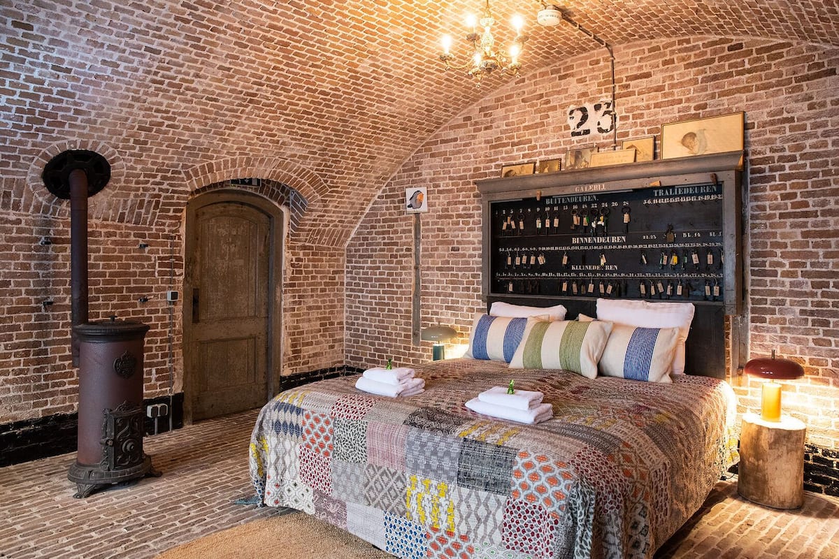 A room with exposed brick walls, a wooden door, a bed with a quilt, a wall clock, and a candle on a side table.