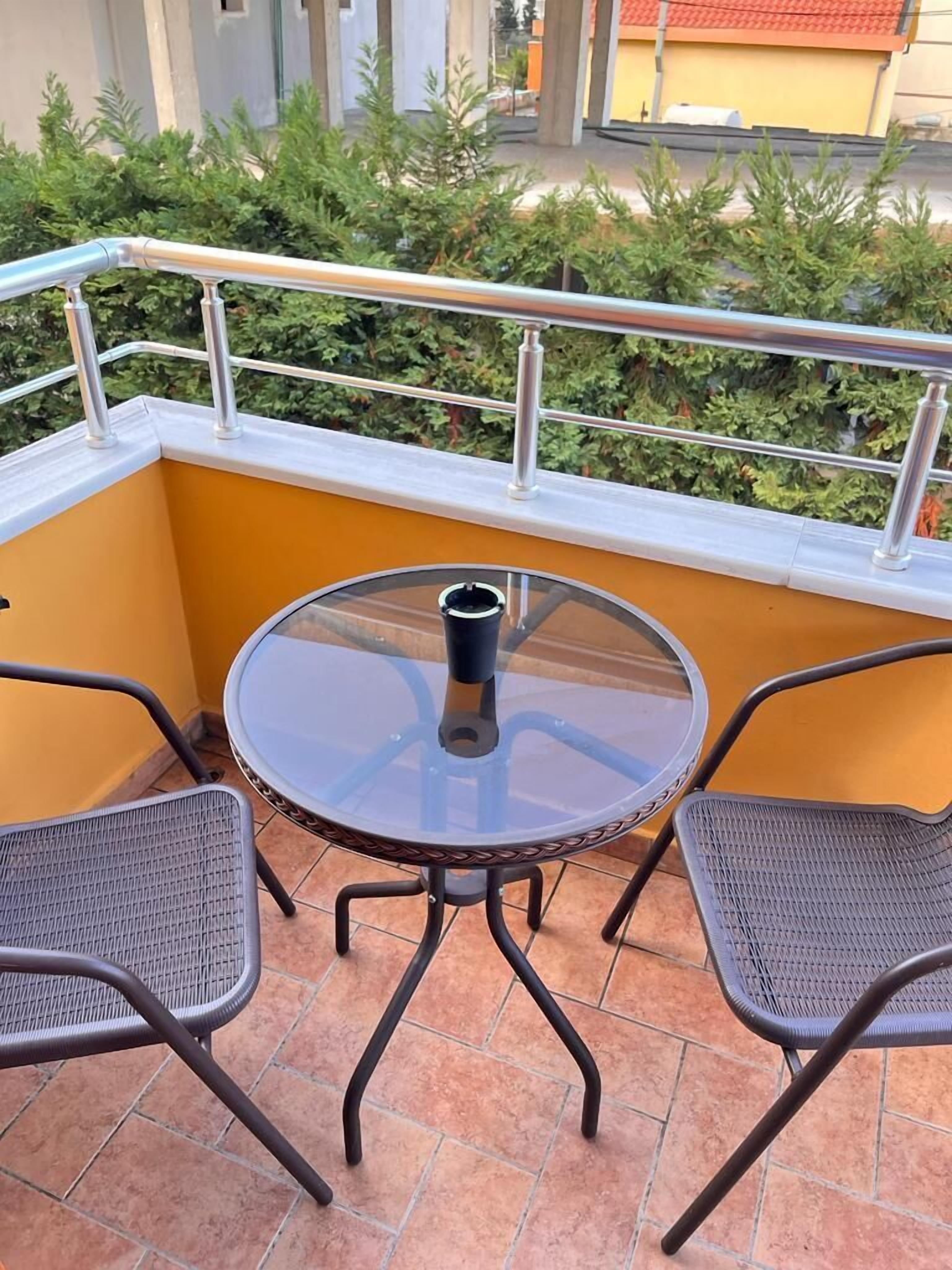 A round glass table with two metal chairs on a balcony with a metal railing.
