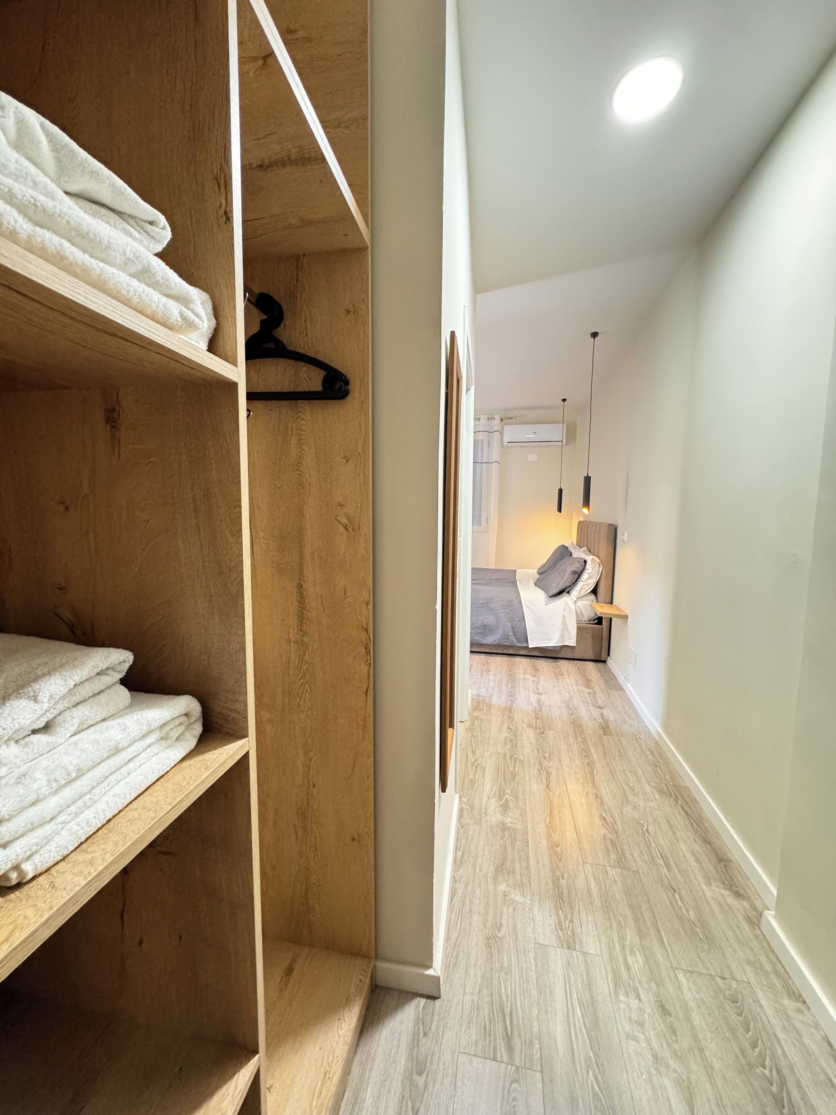 Um corredor estreito com estantes de madeira à esquerda, uma cama com colchão e almofadas cinzentas ao fundo, e uma parede branca com uma luminária no teto.