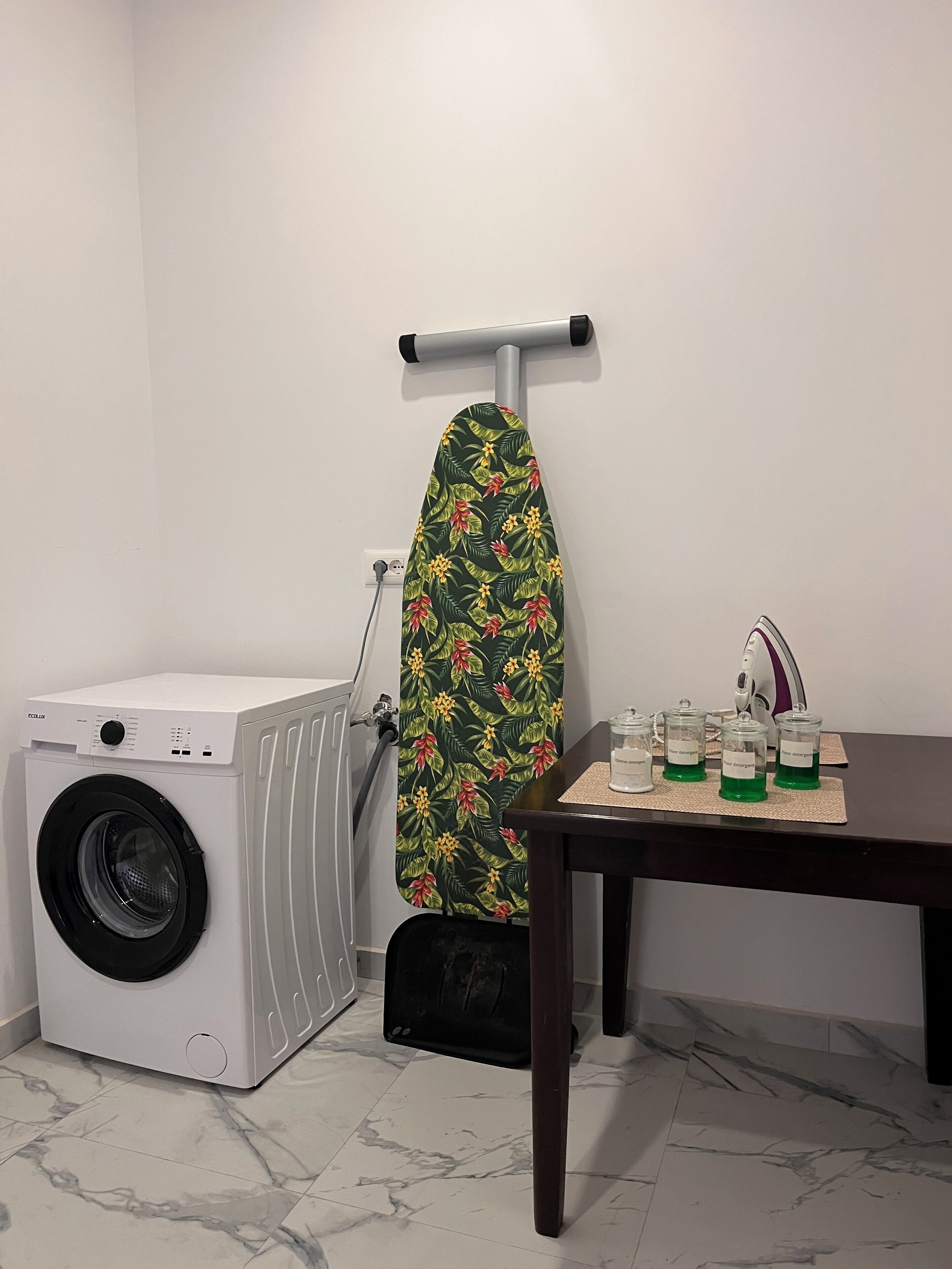 Un espace dédié à la lessive, comprenant un lave-linge, une planche à repasser recouverte d’un tissu à motifs floraux et une table sur laquelle sont posées des bouteilles de lessive.