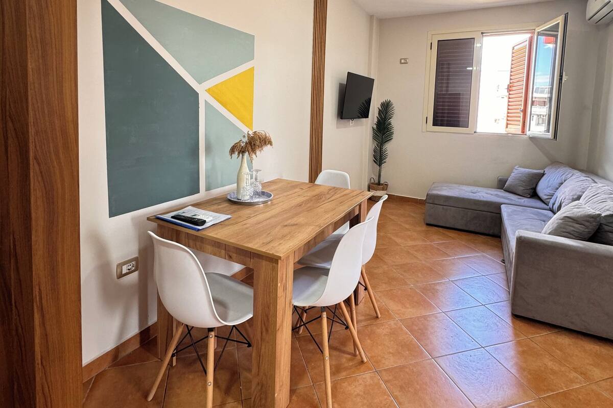 Zona de menjador amb taula de fusta i cadires blanques, un sofà gris i un televisor a la paret.