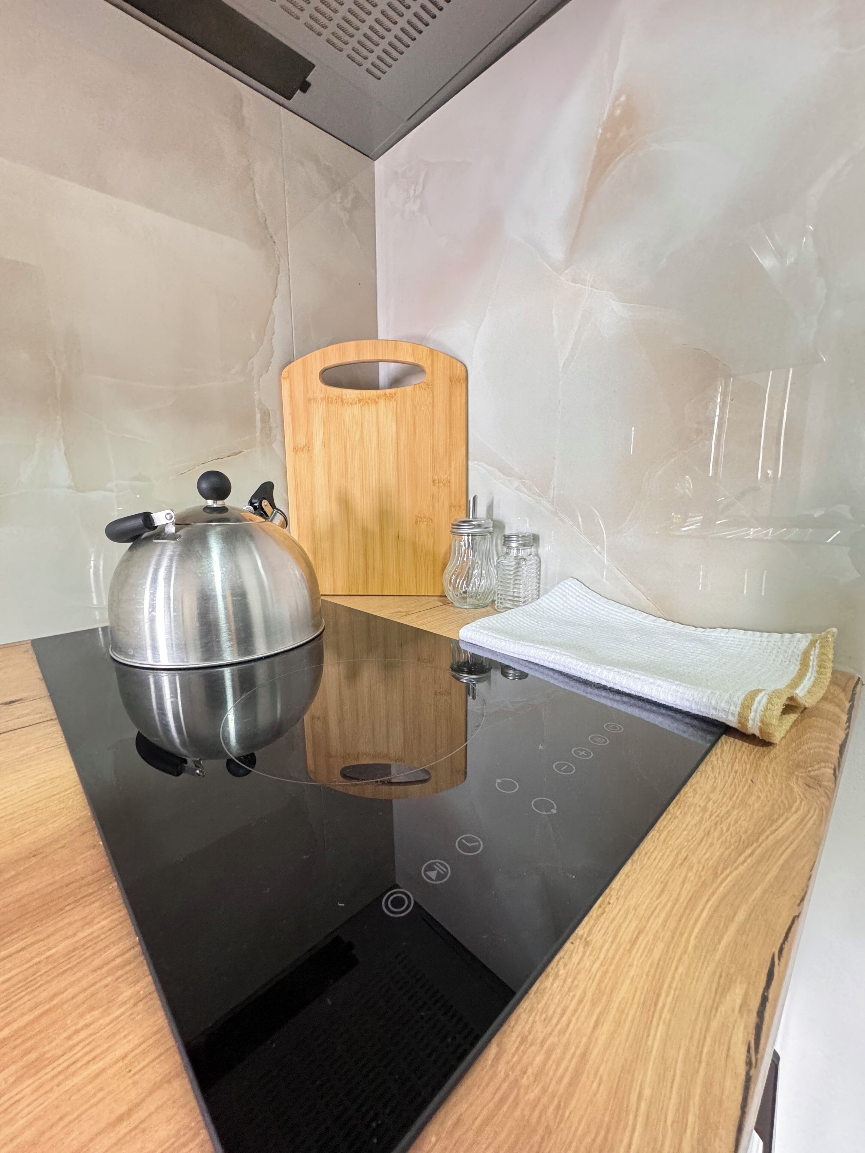A kitchen with a black induction cooktop, a stainless steel kettle, a wooden cutting board, and a white towel.