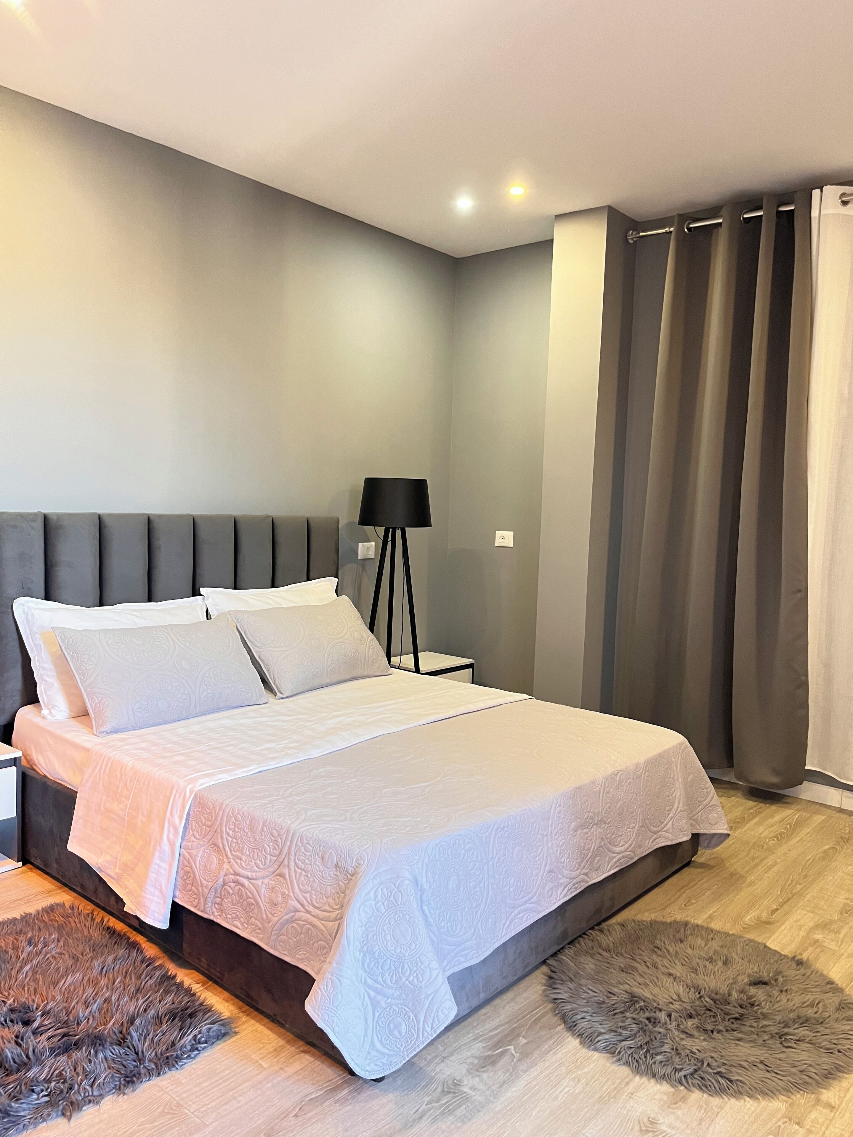 A modern bedroom with a large bed, a bedside table, a floor lamp, and a view of the outside through curtains.