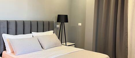 A modern bedroom with a large bed, a bedside table, a floor lamp, and a view of the outside through curtains.