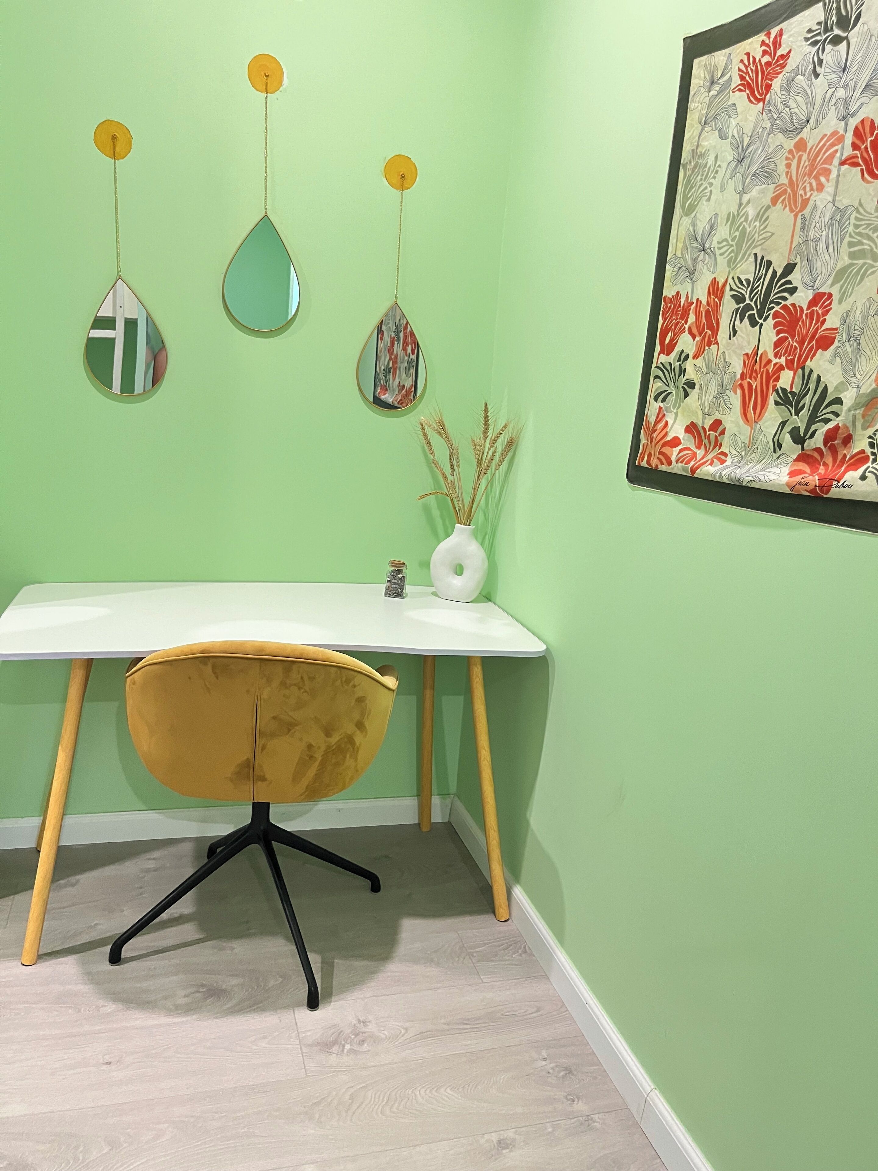 Een hoekappartement met een wit bureau, een gele stoel, een witte vaas met gedroogde planten en een bloemrijk kunstwerk aan de muur.