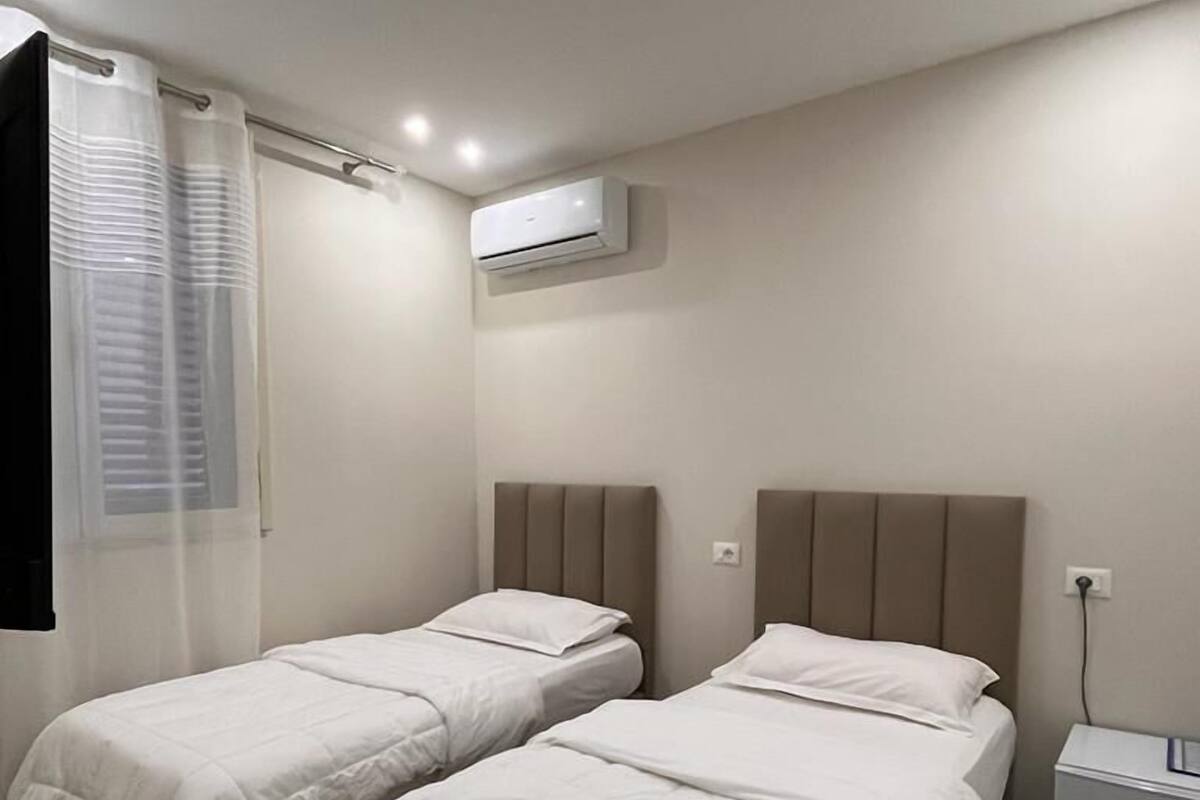Habitació d'hotel amb dos llits individuals, una finestra amb cortina fina, un aparell d'aire condicionat i una taula de nit amb làmpada.